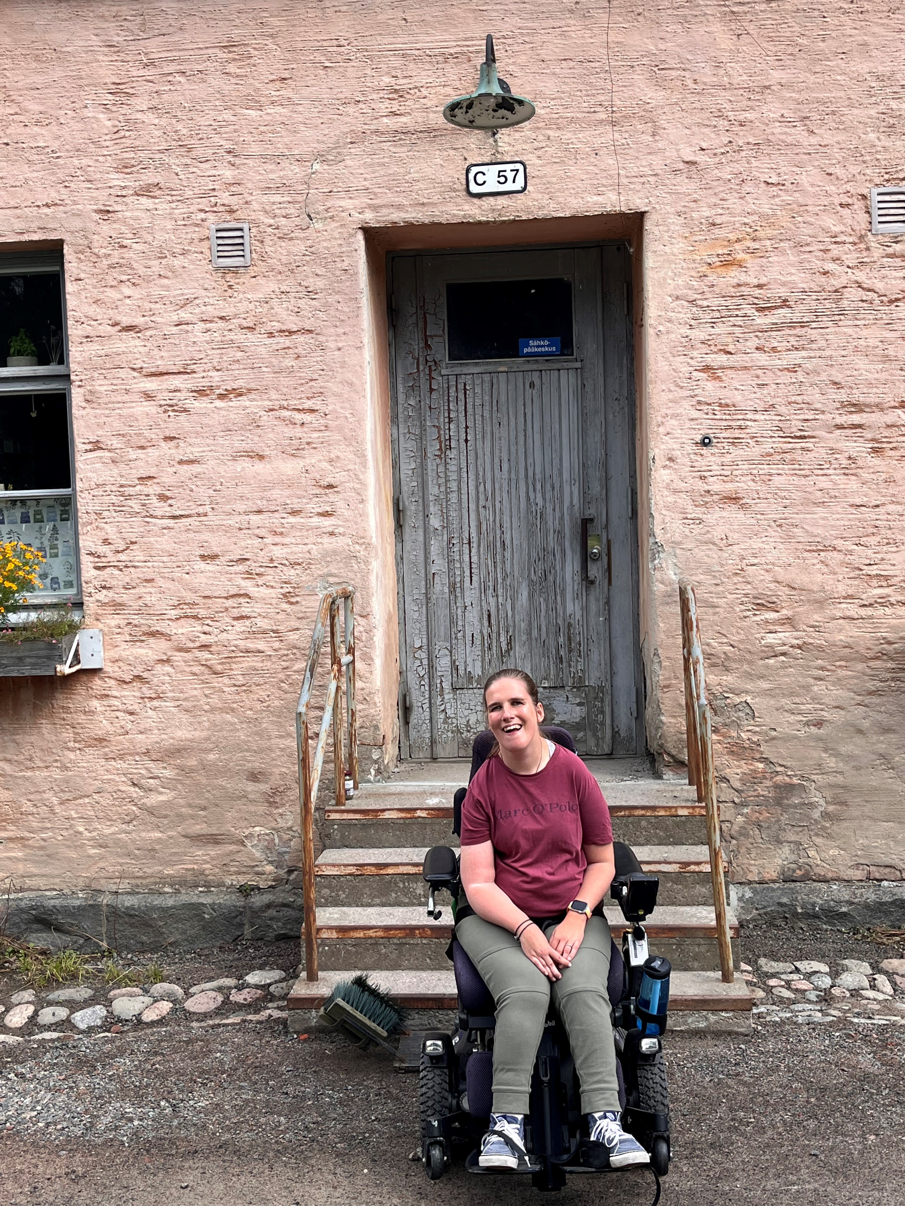 Foto von Lea Voitel, eine junge Frau im Rollstuhl/Das Bild zeigt sie vor einer grauen Tür aus Holz eingebettet in rosafargigem Stein in Suomenlinna, der Festungsinsel bei Helsinki